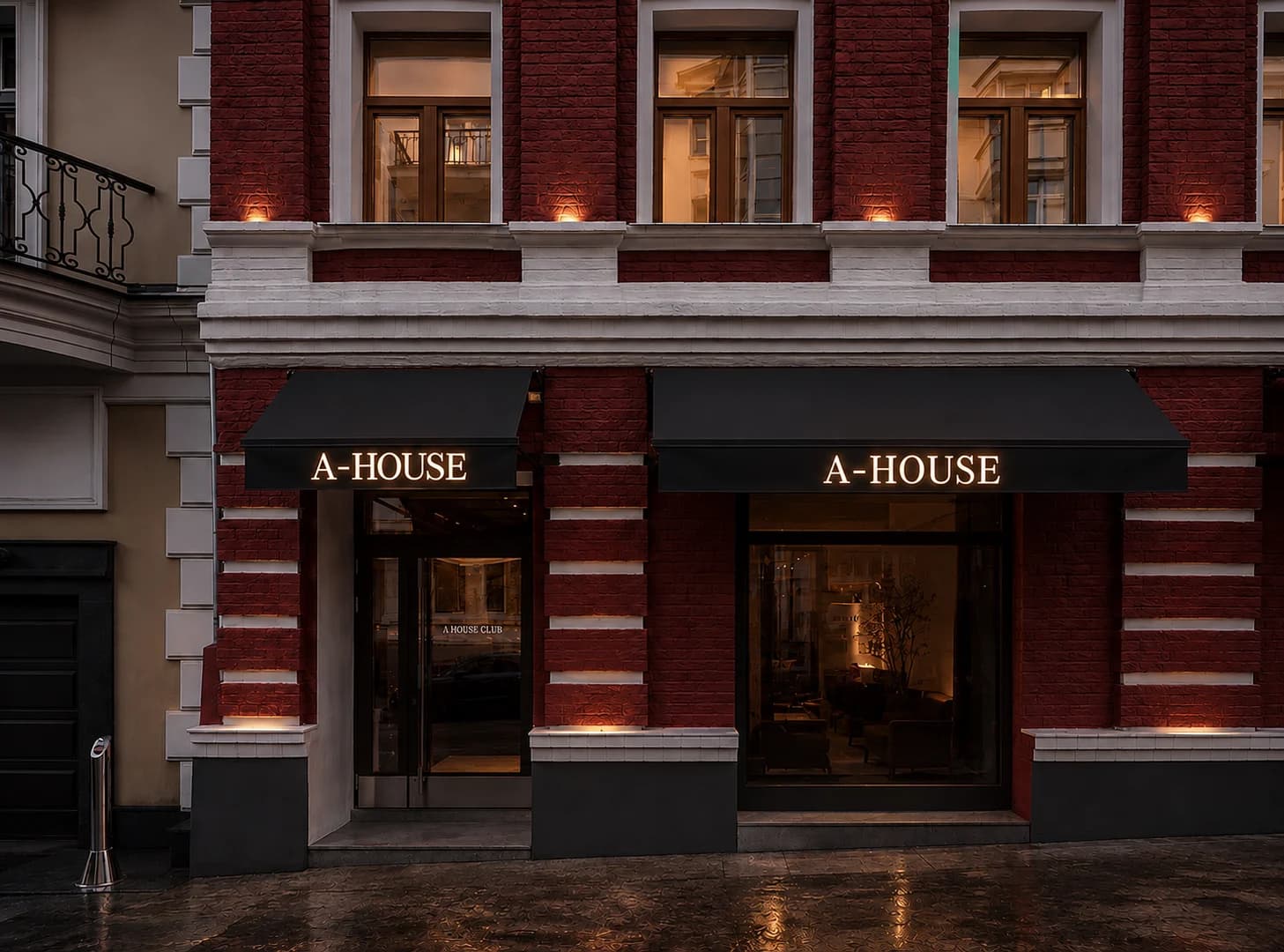 Nächtliche Straßenansicht einer roten Backsteinfassade mit zwei dunklen Markisen und warm leuchtenden Schriftzügen „A-HOUSE“.