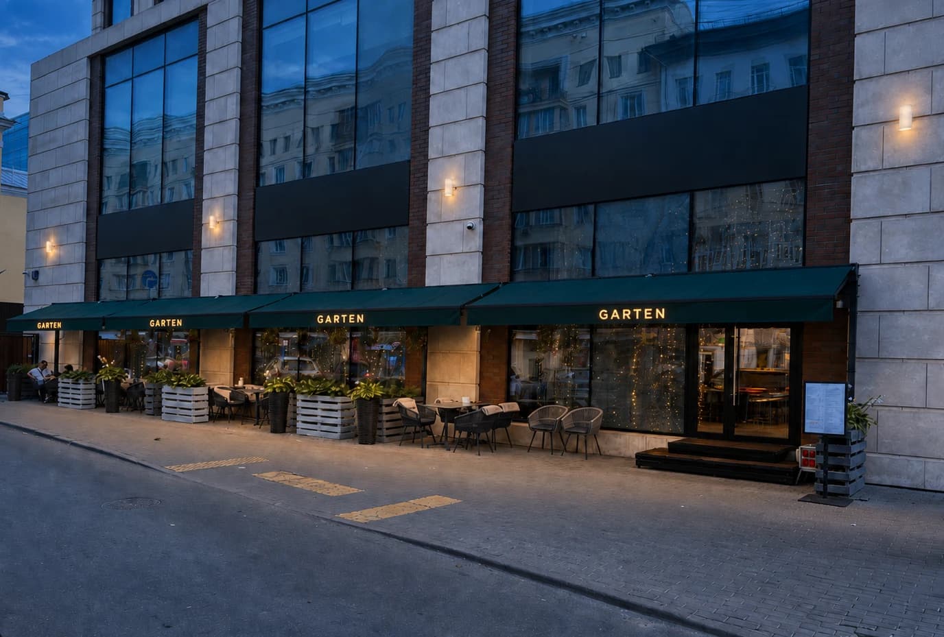 Restaurantfassade mit mehreren dunkelgrünen Markisen und warm leuchtenden Schriftzügen „GARTEN“ über einer Außenterrasse.